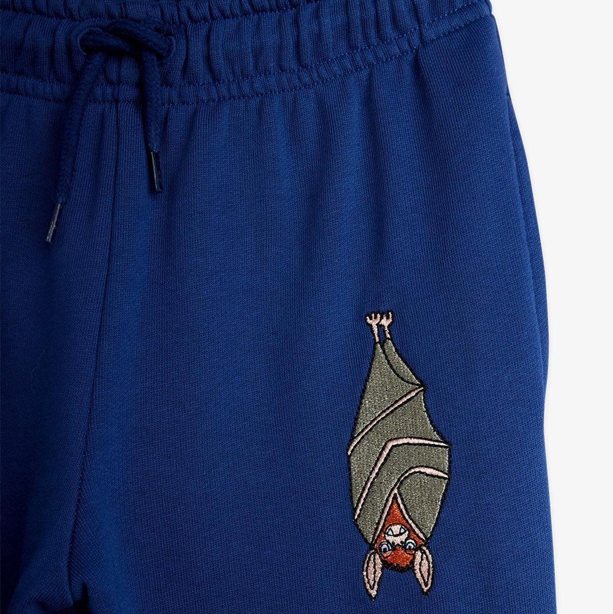 Close-up of a pair of royal blue sweatpants featuring an embroidered bat character on the left leg, with a matching drawstring waistband.