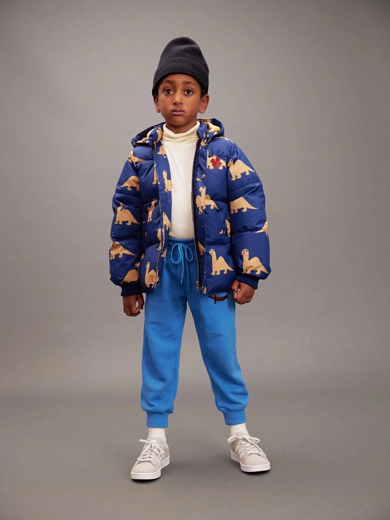 Child wearing a blue jacket with dinosaur pattern and blue pants on a gray background