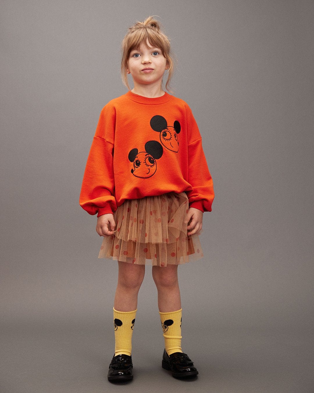 Child wearing an orange sweatshirt with a design, brown tutu skirt, and yellow socks with black mouse ears on a gray background.