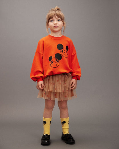Child wearing an orange sweatshirt with a design, brown tutu skirt, and yellow socks with black mouse ears on a gray background.