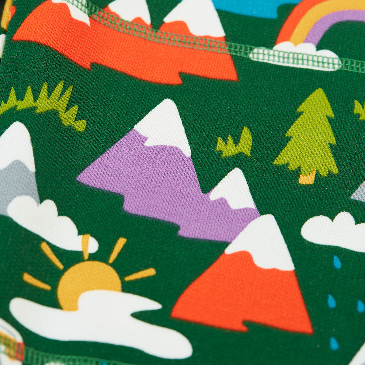 A close-up of a dark green fabric printed with a colorful children's landscape pattern featuring mountains, trees, clouds, a rainbow, and a sun.