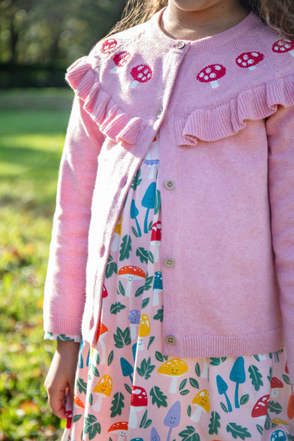 Child wearing a pink cardigan with ruffled details and a colorful dress outdoors.
