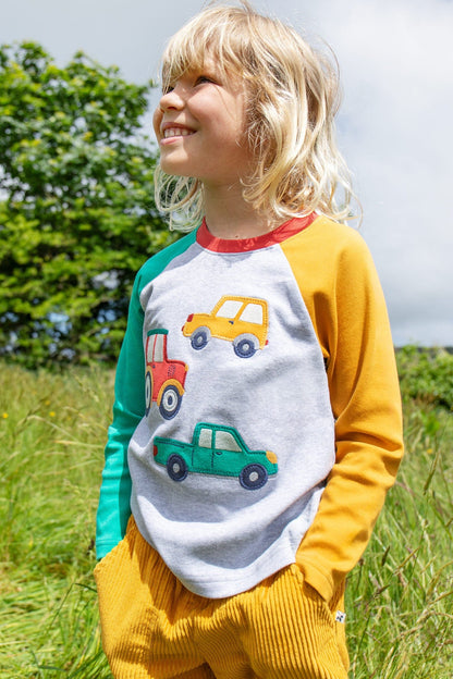 Child wearing Frugi's Alfie vehicles Raglan top with yellow corduroy pants on a field with high grass.