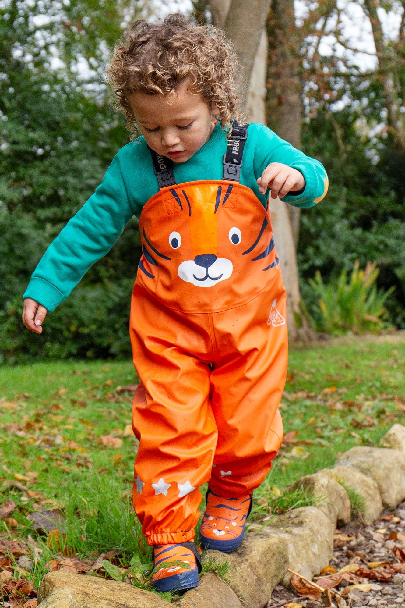 Child wearing Frugi  puddle busters in tiger print outside in the tree, balancing on rocks. Autumn leaves are scattered around on the ground