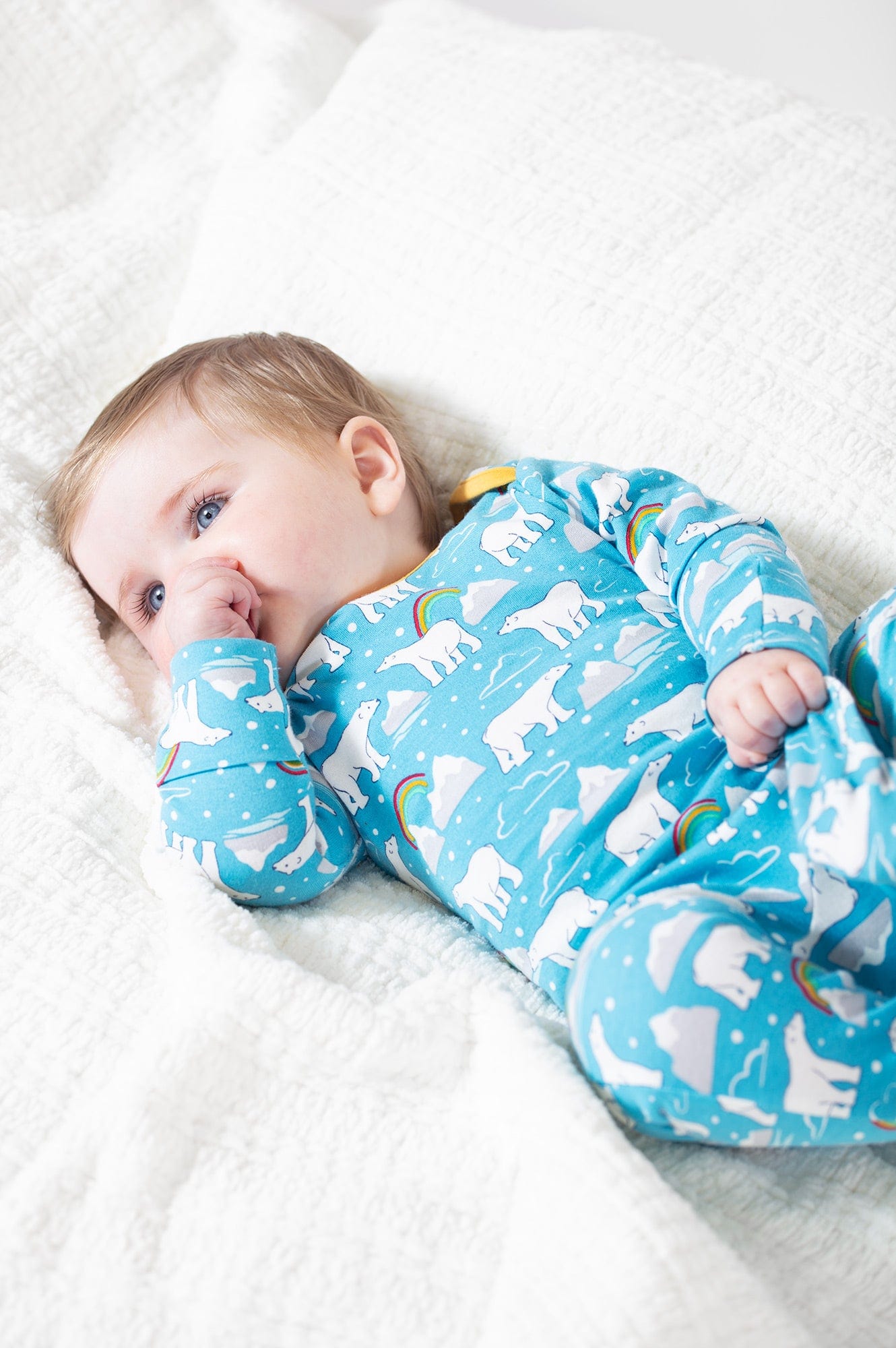 Baby wearing Frugi's baby grow in winter print in light blue, polar bears and rainbows. Lying on a white muslin cloth.