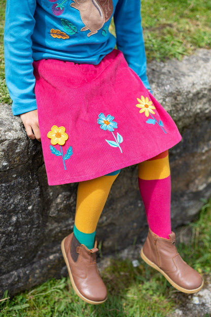 Frugi skirt in corduroy worn with blue top and colour block tights. Child is sitting on a stonewall surrounded by green grass.