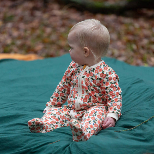 A baby sitting on a teal blanket outdoors, wearing a footed zip-up sleeper with a coral and green floral pattern on a cream background.