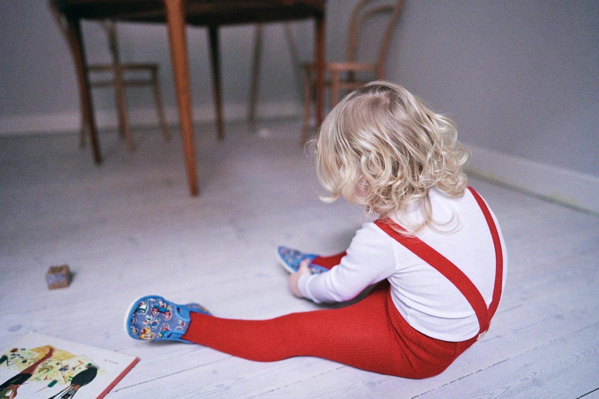Child sitting on the floor with a book and toys in a room with wooden furniture.
