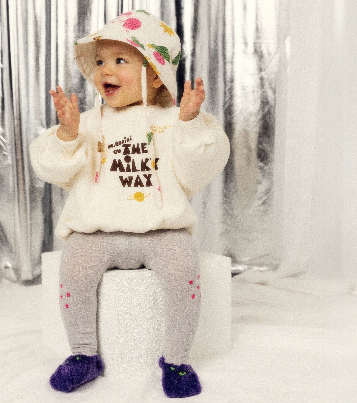 Child wearing a white sweatshirt with text, gray pants, and colorful hat against a silver curtain background.