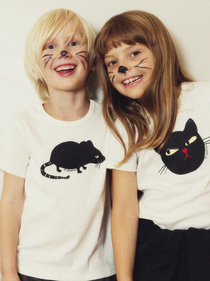 Two children smiling and posing together, wearing white graphic t-shirts with black cat and mouse illustrations and cat face makeup on their cheeks.